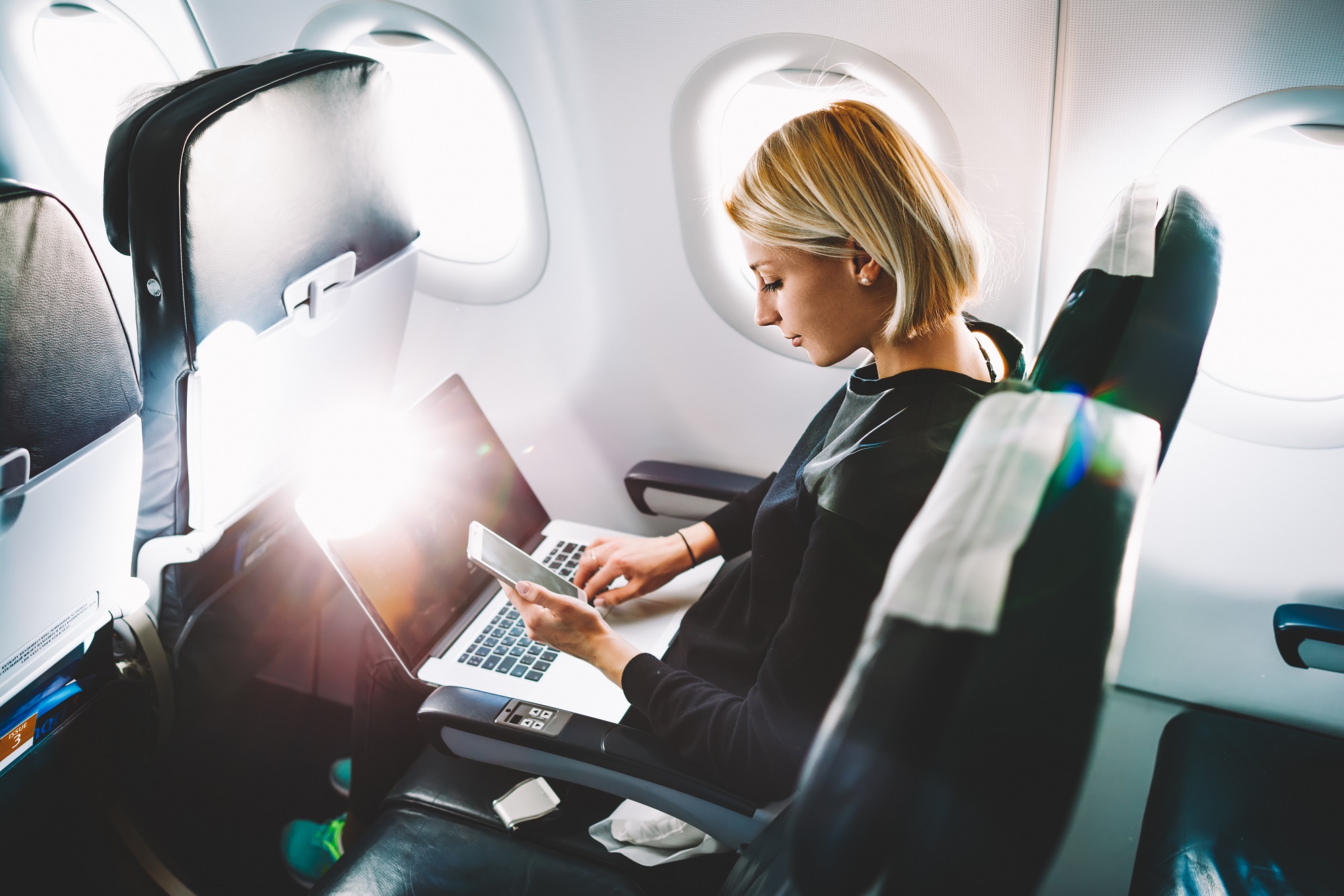 A professional woman is sitting in a seat on an airplane while working on her laptop.
